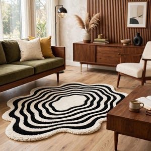 Unique hand-tufted organic wave rug in deep black and ivory white, featuring layered concentric curves and a softly scalloped outer edge. The artistic statement rug adds dramatic contrast to a neutral living room with a cream sofa, wooden coffee table, beige throw, indoor olive tree, minimalist décor and large bright windows.