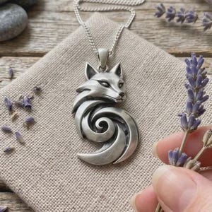 May include: A silver-toned pendant necklace featuring a stylised fox head with a swirling, curved tail design. The pendant hangs from a delicate silver chain, resting on a light beige fabric square. The fox has pointed ears and detailed facial features.