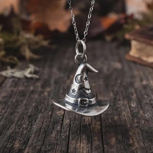 Puede incluir: Un colgante de metal plateado con forma de sombrero puntiagudo, suspendido de una fina cadena a juego. El sombrero presenta detalles en relieve que incluyen una luna creciente, estrellas, un pequeño murciélago y una banda con hebilla.