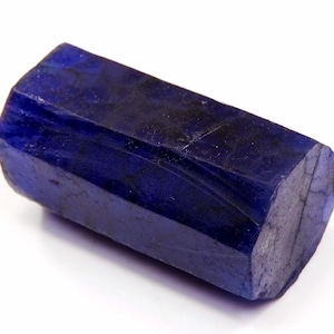 May include: A raw, hexagonal sapphire crystal with a deep, saturated royal blue colour. The stone features a natural, unpolished surface with visible internal fractures and crystalline textures, displaying a distinct elongated, six-sided prism shape.
