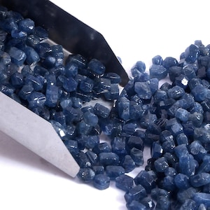 Pode incluir: Uma pá de metal despejando uma grande coleção de pequenas pedras preciosas de safira azul-escuras polidas. As pedras têm formas irregulares com superfícies facetadas, exibindo vários tons de azul-marinho e real, espalhadas sobre uma superfície branca.