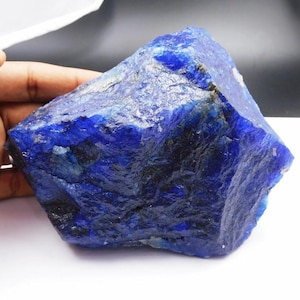 May include: A large, raw, unpolished chunk of deep blue sapphire mineral. The stone features a jagged, crystalline surface with varying shades of intense cobalt and navy blue, showing natural fractures and rough, uneven edges throughout the rock.