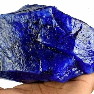 Puede incluir: Una gema de zafiro grande, cruda y sin cortar con un color azul real profundo y vibrante. La piedra presenta una textura cristalina rugosa con facetas naturales, crestas y superficies irregulares que captan la luz, mostrando tonos azules.