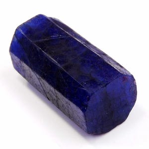 May include: A raw, hexagonal sapphire crystal with a deep, saturated blue colour. The gemstone features a natural, elongated prismatic shape with visible surface textures, internal fractures, and distinct geometric facets along its length.