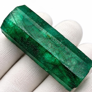 Può includere: Un cristallo di smeraldo grezzo e allungato con una tonalità verde intenso e una struttura prismatica esagonale. La superficie presenta fratture interne naturali, vari livelli di traslucenza e una consistenza ruvida e grezza.