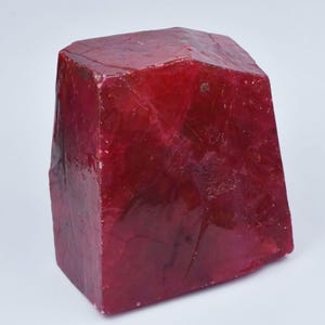 May include: A large, raw ruby crystal with a deep red color and a translucent, multifaceted surface. The stone features a blocky, irregular shape with visible internal fractures and natural textures throughout its solid, crystalline structure.