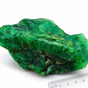 Pode incluir: Uma esmeralda bruta e não polida de cor verde profunda com uma textura cristalina visível. A pedra está posicionada ao lado de uma régua branca, mostrando um comprimento de aproximadamente 5 centímetros, destacando sua forma natural e irregular.
