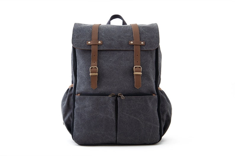 May include: A gray canvas backpack with brown leather straps and buckles. The backpack has a large main compartment and two smaller front pockets.