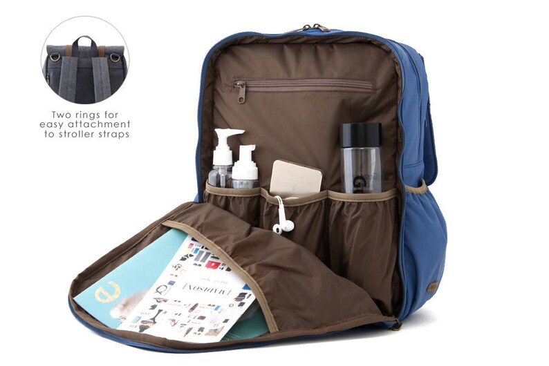 May include: A blue backpack with multiple pockets and compartments. The backpack has a brown interior and features two rings for easy attachment to stroller straps. The backpack is open and shows a magazine, a water bottle, and two bottles of lotion inside.