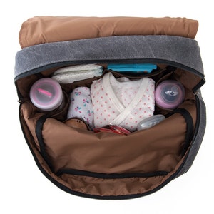 May include: A gray canvas diaper bag with brown accents is open and filled with baby items, including a white onesie with pink polka dots, a blue and white patterned cloth, and two pink bottles.