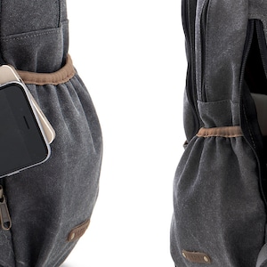 May include: A gray canvas backpack with a zippered pocket on the front. The pocket has a brown leather trim and a zipper pull. A cell phone is visible inside the pocket.