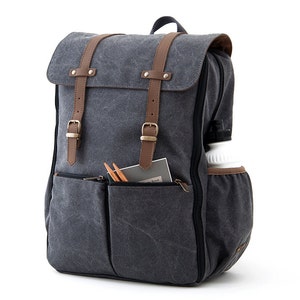 May include: A gray canvas backpack with brown leather straps and buckles. The backpack has a large main compartment and a smaller front pocket. The backpack is water-resistant.