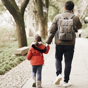 May include: A father and daughter walk hand-in-hand down a paved path. The father is wearing a brown jacket and a gray backpack. The daughter is wearing a red jacket and black leggings. They are walking through a park with trees on either side of the path.