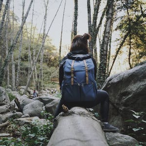 May include: A person wearing a blue backpack sits on a log in a forest. The person is looking out at the trees. There are large rocks in the background.
