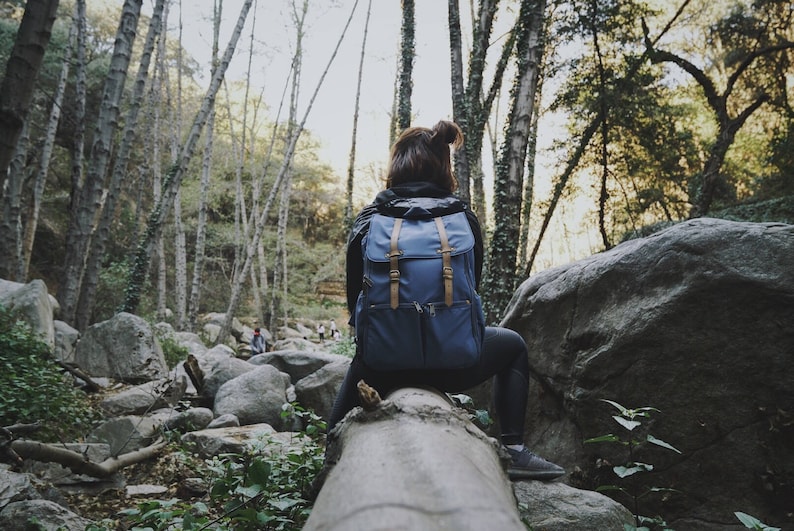 May include: A person wearing a blue backpack sits on a log in a forest. The person is looking out at the trees. There are large rocks in the background.