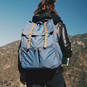 May include: A person wearing a black jacket and blue jeans is carrying a blue backpack with brown straps. The backpack has two zippered pockets on the front.