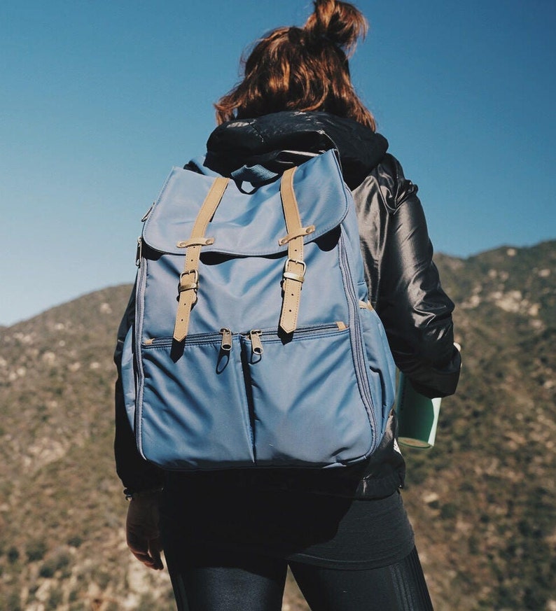 May include: A person wearing a black jacket and blue jeans is carrying a blue backpack with brown straps. The backpack has two zippered pockets on the front.