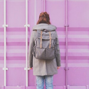 May include: A person wearing a gray coat with a hood and a gray backpack stands in front of a bright pink wall. The person is wearing blue jeans and black shoes.