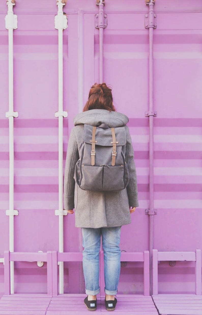 May include: A person wearing a gray coat with a hood and a gray backpack stands in front of a bright pink wall. The person is wearing blue jeans and black shoes.