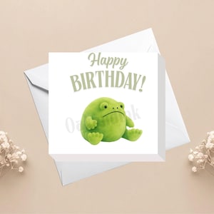 May include: A square white greeting card featuring the text Happy BIRTHDAY in sage green letters above a round, bright green cartoon frog sitting down. The card is positioned on top of a white envelope, accompanied by sprigs of white baby's breath.