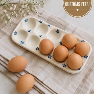 May include: A rectangular ceramic egg tray with a cream-colored glaze and small blue flower patterns. The tray features eight circular indentations, with six brown eggs resting inside the tray and two additional brown eggs placed nearby.