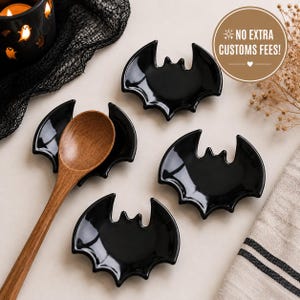 May include: Three black ceramic bat-shaped dishes and one wooden spoon. The dishes feature a glossy finish and a distinct bat silhouette with pointed ears and scalloped wings. A brown wooden spoon rests inside one of the bat-shaped dishes.