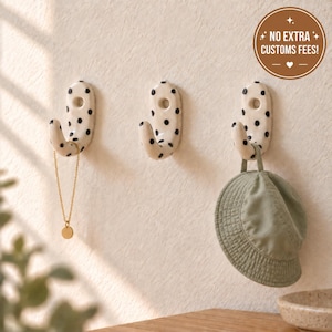 May include: Three off-white ceramic wall hooks with black polka dots. Each hook features a rounded J-shape and a circular mounting hole. A gold chain hangs from the left hook, and a sage green bucket hat hangs from the right hook.