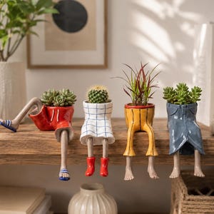Puede incluir: Cuatro macetas de cerámica con forma de parte inferior del cuerpo vistiendo diferentes prendas: pantalones cortos rojos con sandalias azules, falda blanca con cuadrícula azul y botas rojas, pantalones amarillos y falda de mezclilla azul.