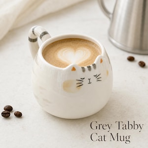 May include: A white ceramic mug shaped like a cat with grey tabby stripes, featuring a handle designed as a striped tail, small ears on the rim, and a painted face with whiskers. The mug is filled with coffee featuring heart-shaped latte art.