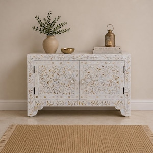 Peut inclure: Un buffet rectangulaire blanc orné de motifs floraux complexes en nacre sur deux portes, doté de deux boutons en cristal transparent et d'une base solide. Le dessus accueille un vase beige avec de la verdure, un petit bol doré et une lanterne.