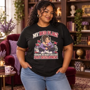 May include: A black short-sleeve t-shirt featuring a graphic of a person reading a book surrounded by red flags. The text reads: MY RED FLAG? THINKING HIS RED FLAGS ARE PLOT DEVELOPMENT. The design includes a mug, books, and sparkles.