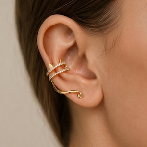 May include: A gold-toned ear cuff featuring a cat-ear design with two parallel bands encrusted with small clear stones. The piece extends into a smooth, curved tail-like shape that follows the contour of the ear cartilage and lobe.