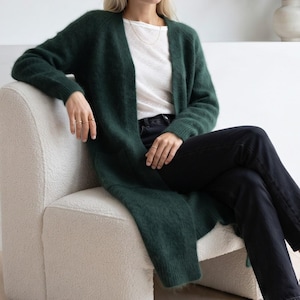 May include: A long-sleeved, open-front cardigan in a deep forest green color with a fuzzy, textured knit finish. The garment features ribbed cuffs and a simple, collarless design, worn over a plain white t-shirt and dark black denim jeans.