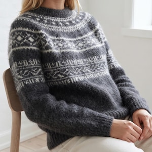 Könnte beinhalten: Ein langärmeliger Pullover in Anthrazit mit flauschiger, gebürsteter Textur und einem weißen geometrischen Fair-Isle-Muster an der Passe und im Brustbereich. Das Kleidungsstück hat einen Rundhalsausschnitt sowie gerippte Bündchen an Ärmeln und Saum.