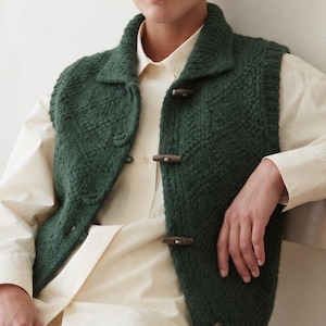 May include: A dark green sleeveless knit vest featuring a textured diamond pattern, a wide collar, and three brown toggle closures down the front. The garment is worn over a light cream-colored button-down shirt with long sleeves and a collar.