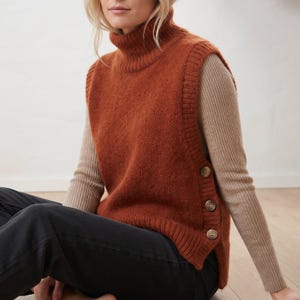 May include: A rust-coloured sleeveless knit sweater vest featuring a high turtleneck collar and a ribbed hem. The side of the garment is detailed with three round, light-coloured buttons arranged vertically along the seam, layered over a beige long-sleeve top.