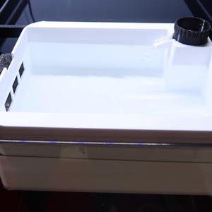 Puede incluir: Una caja de filtro de acuario de plástico blanco con una tapa circular negra y un inserto de espuma negra. La unidad presenta un diseño de cubeta rectangular con un pequeño pico y está colocada dentro de una estructura de tanque de vidrio. Logo VIREX AQUATICS.