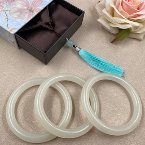 May include: Three smooth, translucent white jade-style bangles arranged in a cluster. A dark brown gift box with a light blue floral lid sits nearby, accompanied by a bright turquoise tassel and a single pale pink artificial rose flower.