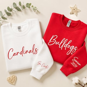 May include: A white sweatshirt with red text reading Cardinals and Go Cards on the sleeve, alongside a red sweatshirt with white text reading Bulldogs and Senior 2024 on the sleeve. Both garments feature crew necklines and long sleeves.