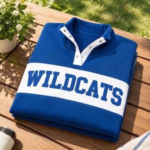 Op de afbeelding: Een koningsblauw sweatshirt met een witte horizontale borststreep waarop het woord WILDCATS in blauwe blokletters is gedrukt. Het kledingstuk heeft een witte sluiting met zilverkleurige metalen drukknopen bij de kraag.