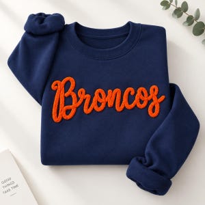 Può includere: Una felpa girocollo blu navy con la scritta Broncos in un carattere corsivo arancione testurizzato e in rilievo sul petto. Il capo presenta maniche lunghe con polsini a coste e una classica scollatura rotonda.