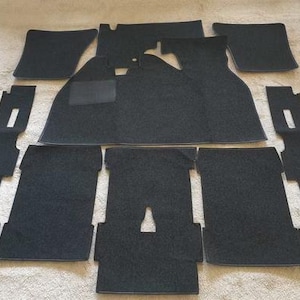 Puede incluir: Un juego de alfombrillas negras para automóvil con una superficie de moqueta texturizada. La colección incluye varias piezas con recortes y formas específicas diseñadas para el interior del vehículo, incluyendo una alfombrilla principal.