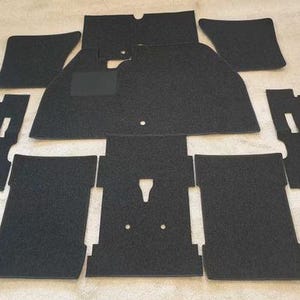 May include: A set of black automotive floor mats consisting of multiple pieces with specific cutouts and shapes. The mats feature a textured surface and include a rectangular heel pad on the driver side piece, designed for vehicle interior coverage.
