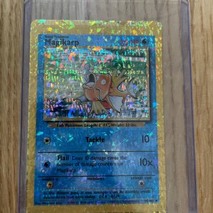 Peut inclure: Une carte à collectionner holographique Magikarp dans une pochette de protection en plastique transparent. La carte présente une bordure dorée sur fond bleu, illustrant le personnage, le nom Magikarp, 30 PV et les attaques Charge et Fléau.