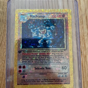 Può includere: Una carta collezionabile olografica Machamp in una custodia protettiva in plastica trasparente. La carta presenta il personaggio blu a quattro braccia, il testo Machamp, 100 HP, Seismic Toss 60 e un timbro 1st Edition su sfondo dorato.