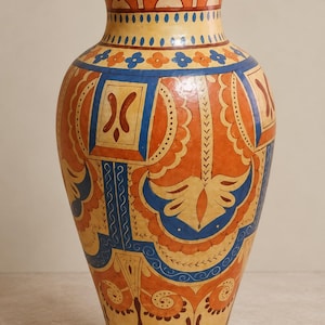 Puede incluir: Un jarrón de cerámica alto con un diseño vibrante pintado a mano en tonos naranja, azul y crema. El cuello tiene un borde verde sólido, mientras que el cuerpo muestra intrincados patrones geométricos, motivos florales y volutas.