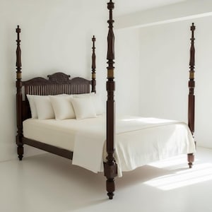 Op de afbeelding: Een donkerbruin houten hemelbed met hoge, gedraaide palen en een hoofdeinde met verticale latten en een uitgesneden centraal ornament. Het bed is opgemaakt met een effen wit hoeslaken, een laken en vier bijpassende witte vierkante kussens.