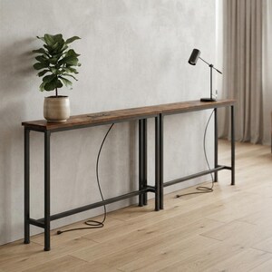May include: A long, narrow console table with a dark wood-grain top and a black metal frame. The table features a built-in power outlet on the surface. A potted green plant sits on the left side, and a black desk lamp is positioned on the right side.