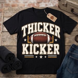 May include: A black short-sleeve t-shirt featuring a graphic print of an American football centred between the words THICKER and KICKER in a distressed white font. The design includes a yellow goal post outline and small white stars.