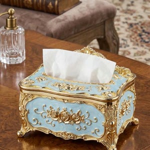 May include: A rectangular tissue box holder featuring a light blue enamel finish with ornate gold-tone metal filigree detailing. The design includes raised gold floral motifs on the sides and four decorative feet, holding a stack of white tissues.